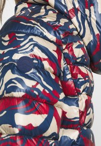 Donsjack in marineblauw, crème en rood zebrapatroon. Heeft doorgestikte delen, een glanzende textuur en een ronde logopatch op de mouw.