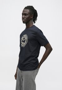 Tumesinine puuvillane t-särk, mille rinnal on ümmargune kreemjas graafiline logo. Ümar kaelus, lühikesed varrukad ja lõdvestunud istuvus.