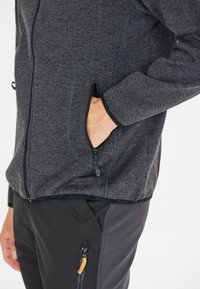 Grå zip-up jakke med tekstureret stof, der har en sidelomme, kontrastfarvet sort kant og en tætsiddende pasform.