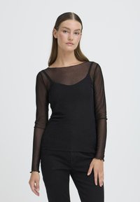 Maglietta nera a maniche lunghe con overlay trasparente a pois, scollatura a barca e sottogiacca aderente, arricchita da leggere ruches sui polsini.