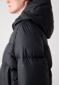 Personne portant une veste matelassée noire avec capuche, vue de profil des épaules vers le haut sur un fond uni.