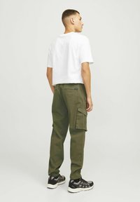 Pantaloni cargo verde oliv cu buzunare laterale, manșete elastice și un patch negru cu logo, asortați cu un tricou alb simplu și adidași negri.