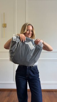 Graues Kleidungsstück, verpackt in klarem Plastic, mit einer glatten Textur. Etiketten sind sichtbar. Das Modell trägt dunkelblaue Jeans in einem hellen Innenraum.