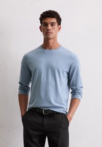 Himmelblaues Langarmshirt aus weichem Stoff mit Rundhalsausschnitt, kombiniert mit schwarzen Hosen und einem dunklen Gürtel. Dezente Texturdetails sichtbar.