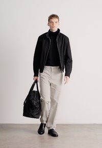 Μαύρο σακάκι με φερμουάρ πάνω από μαύρο ζιβάγκο, μπεζ παντελόνι κατ' ευθείαν και μαύρα παπούτσια. Κρατάει μια μαύρη τσάντα duffle, σε ουδέτερο φόντο.