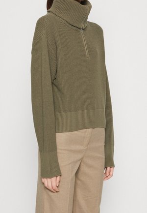 Pull à col roulé en tricot vert olive avec demi-fermeture éclair, porté avec un pantalon beige ajusté sur une personne debout.