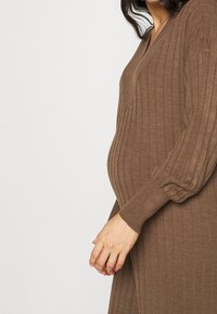 Persona incinta che indossa un vestito in maglia a coste marrone, con le mani posate delicatamente davanti al ventre arrotondato su uno sfondo neutro.