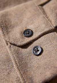 Bruin visgraatstof met twee zwarte knopen met vier gaten. Gedetailleerd stiksel en een kraag zijn zichtbaar in het gestructureerde materiaal.
