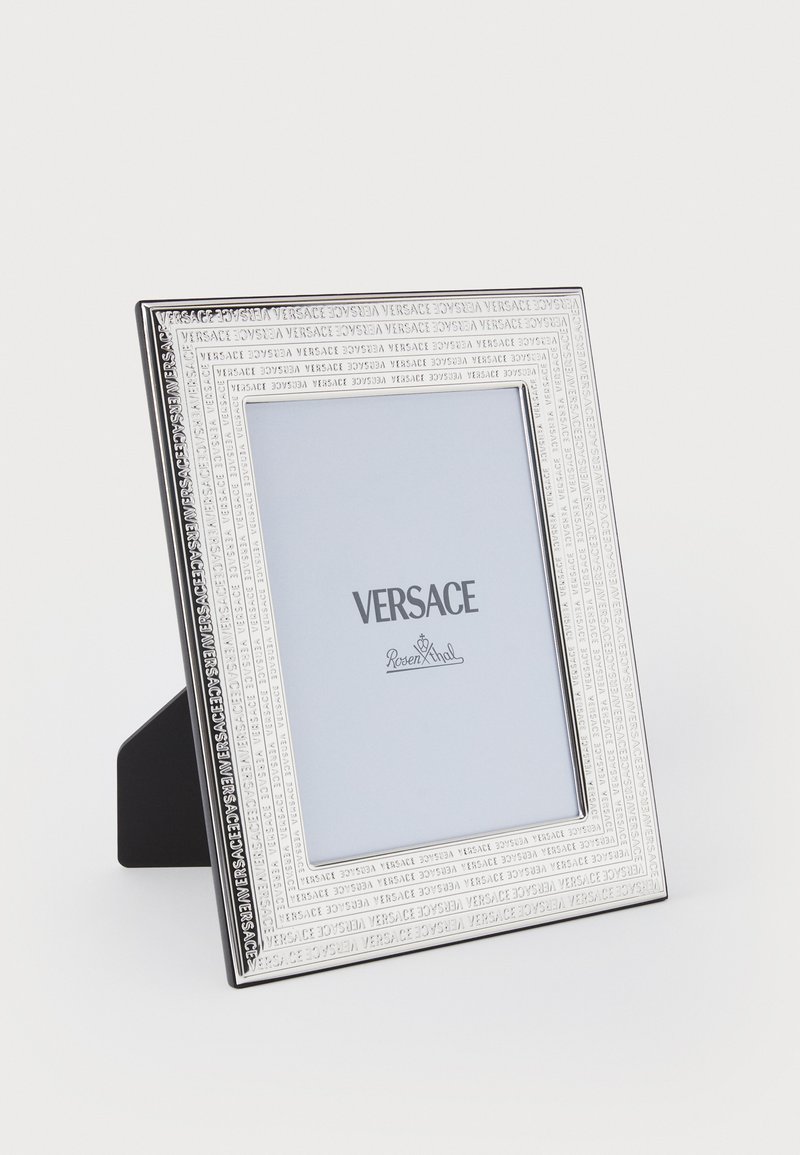 Rektangulär silverram med präglad kant där namnet "Versace" syns, stödd av ett svart ställ och med en vit bakgrund.