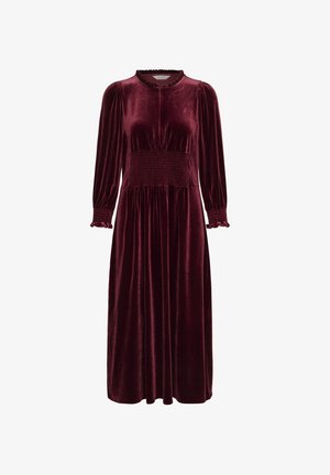 Burgundy velvet dress with long sleeves, smocked waist, ruffled cuffs, and a high neckline. Features a flowing design and soft texture.