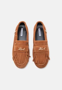 Καφέ σουέντ μοκασίνια με φύλλο σχεδίασης, χρυσές λεπτομέρειες και λογότυπο στον ιμάντα. Λείο εσωτερικό και επίπεδη σόλα.