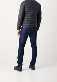 Svart dunjacka med horisontell quiltad struktur, kombinerad med mörkblå jeans och svarta skor, sedd bakifrån.