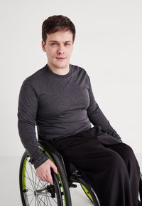 Fekete kerekesszék zöld kiemelésekkel. Embert látunk, aki egy testhezálló szürke hosszú ujjas pólót és laza fekete nadrágot visel, kényelmesen ül.