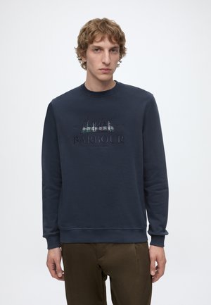 Homme aux cheveux bouclés portant un sweat-shirt bleu marine avec un motif forêt et le logo Barbour, associé à un pantalon marron sur fond blanc.