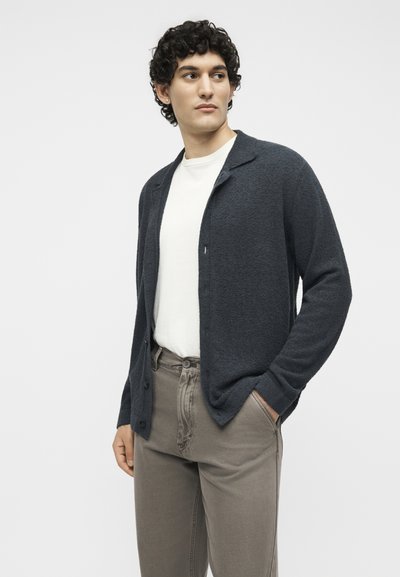 Νέος άνδρας με σγουρά μαλλιά που φοράει σκούρο μπλε ζακέτα πάνω από λευκό πουκάμισο και γκρι παντελόνι, στέκεται με το ένα χέρι στην τσέπη.