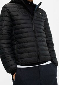 Svart vadderad jacka med quiltad design, huva och dragkedja framtill. Har ribbade muddar och midjeband samt en logotyp på bröstet.