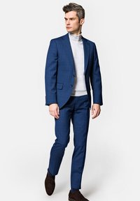 Granatowo-niebieski garnitur w kratkę o dopasowanym kroju, zestawiony z jasnoszarym golfem. Uzupełniony ciemnobrązowymi butami i czystymi liniami.