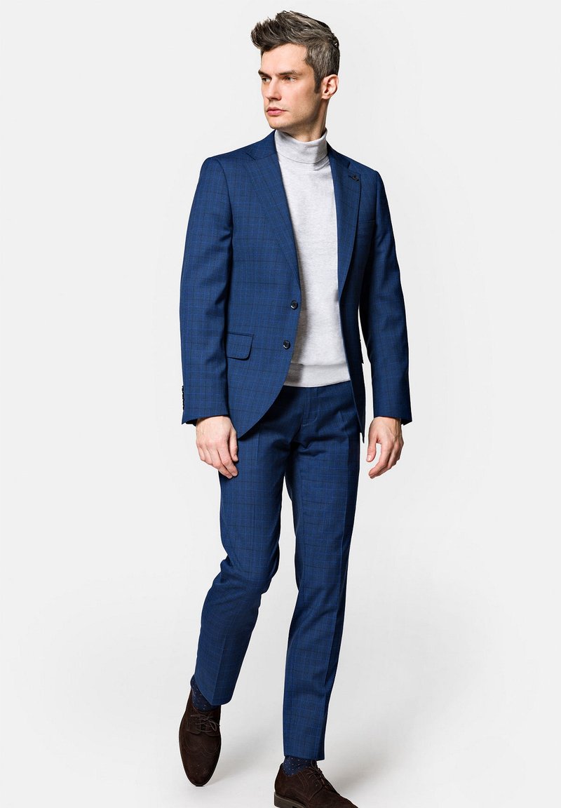 Granatowo-niebieski garnitur w kratkę o dopasowanym kroju, zestawiony z jasnoszarym golfem. Uzupełniony ciemnobrązowymi butami i czystymi liniami.