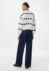 En vit stickad tröja med svarta hjärtformade mönster, långa ärmar och ribbade detaljer, kombinerad med vida mörkblå jeans och bruna skor.