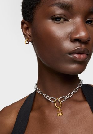 Collana in argento con chiusura e pendente dorato a forma di fiore con accenti rossi; indossata con un piccolo orecchino a cerchio dorato.