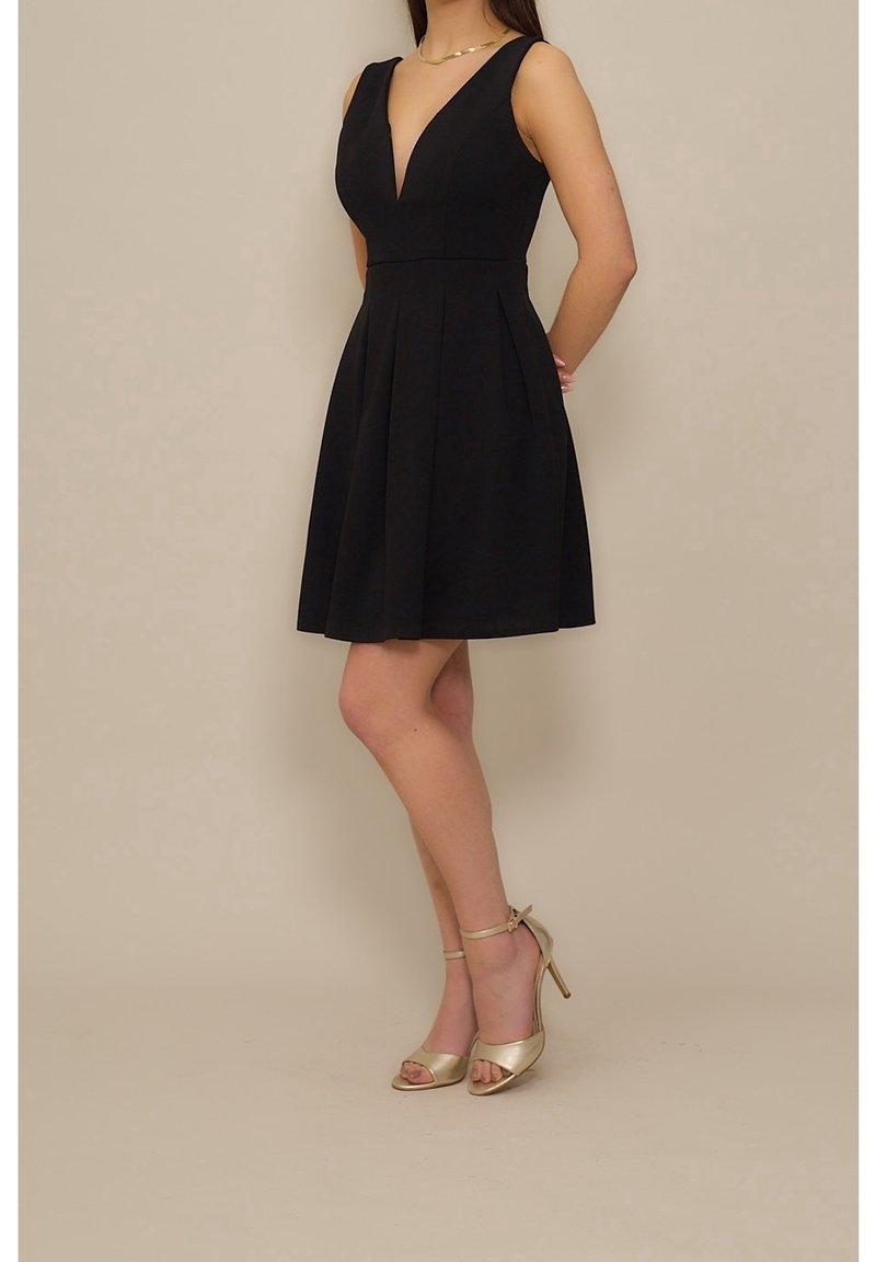 Femme portant une robe noire sans manches avec un décolleté en V profond et des sandales dorées à talons hauts avec bride à la cheville, debout de côté.