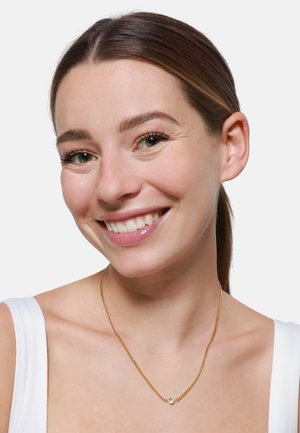 Gouden ketting met een klein, helder rond hangertje, gedragen op een bloot decolleté. Gladde textuur met een glanzende afwerking.