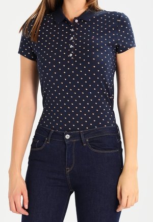 Woman wearing a navy blue polo shirt with a small white and red pattern paired with dark blue jeans, standing with arms relaxed.