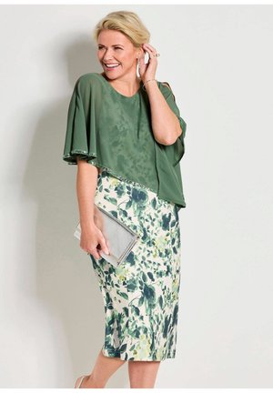 Lächelnde Frau in einem grünen Blumenkleid mit einem transparenten grünen Cape, die eine silberne Clutch hält und ihr Ohrläppchen berührt.