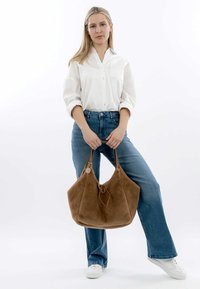 Sac en cuir suédé marron avec une texture douce, deux longues anses et une fermeture à cordon, tenu par une personne portant une chemise blanche et un jean bleu.