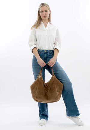 Sac en cuir suédé marron avec une texture douce, deux longues anses et une fermeture à cordon, tenu par une personne portant une chemise blanche et un jean bleu.