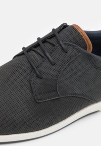 Chaussure perforée noire avec des lacets sombres et un accent en cuir marron près du col. Présente une semelle blanche et une surface supérieure texturée.