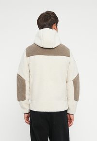 Fleece jacket in cream and brown tones with a hood, featuring textured patches on shoulders and sleeves. Simple, casual design.