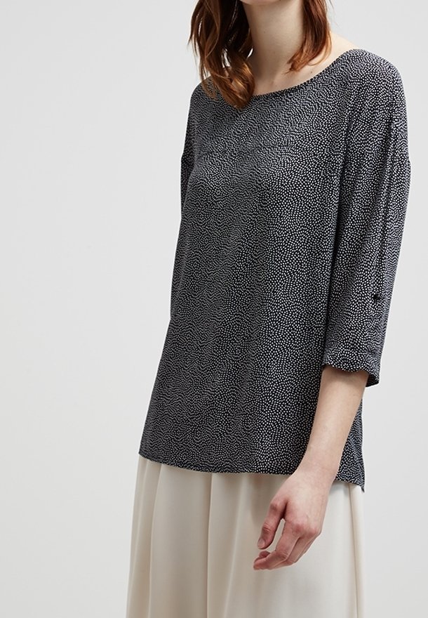 Femme portant un haut ample gris foncé à petits pois blancs et manches trois-quarts, associé à un pantalon beige clair à jambes larges.