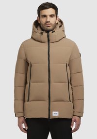 Man in een beige gewatteerde jas met capuchon, met een rits aan de voorkant en ritszakken, staand tegen een effen lichte achtergrond.