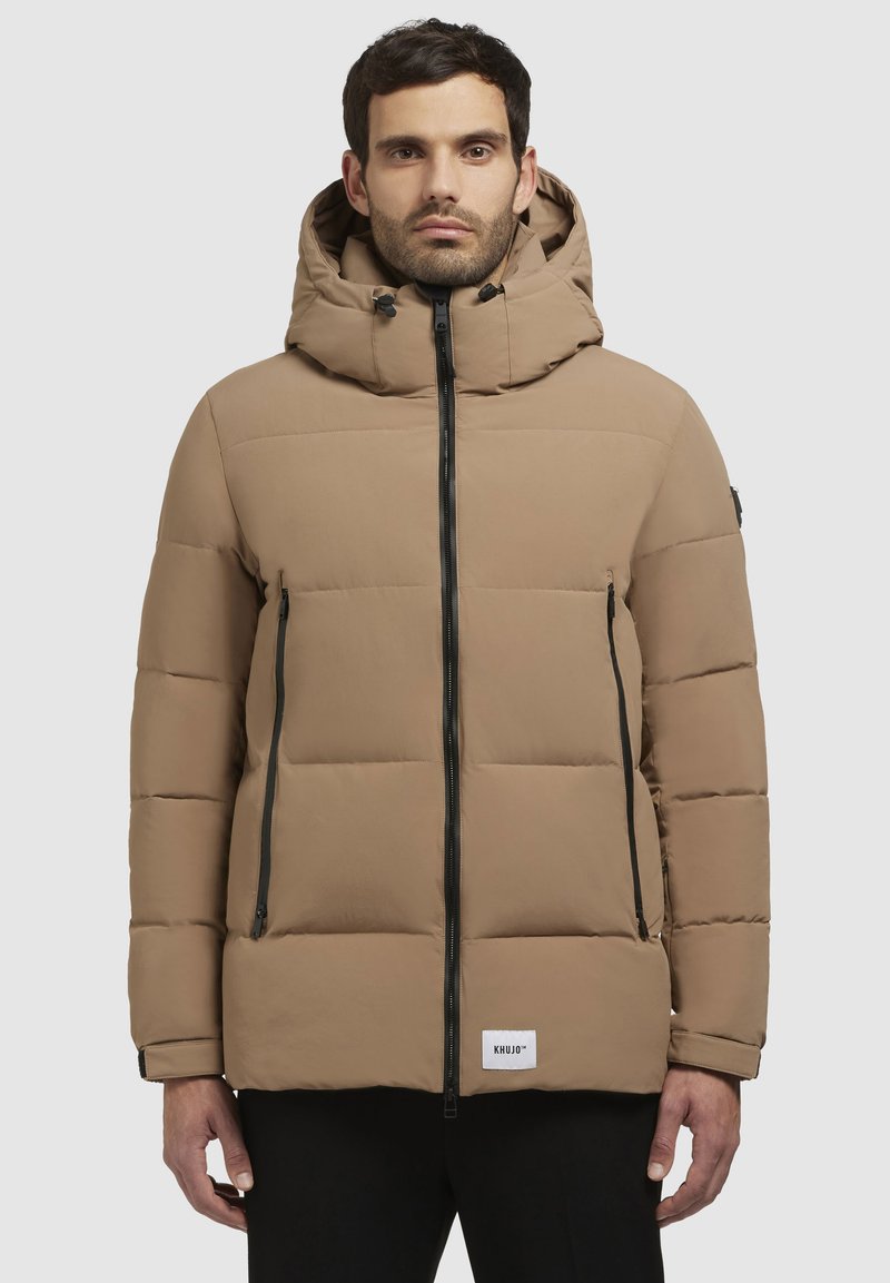 Man in een beige gewatteerde jas met capuchon, met een rits aan de voorkant en ritszakken, staand tegen een effen lichte achtergrond.