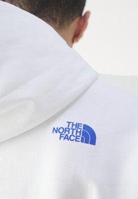 Sudadera blanca con un logo azul en el lado izquierdo, hecha de un tejido suave con un acabado texturizado y capucha con cordón.