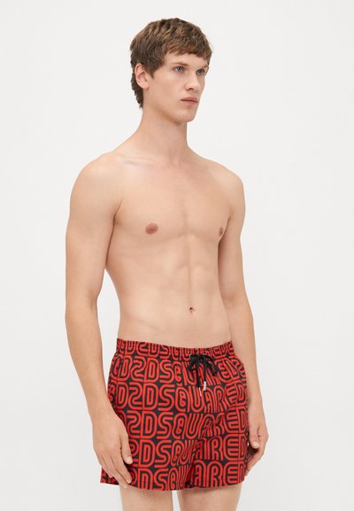 Homme avec des cheveux bruns courts portant un short de bain à motifs rouges et noirs avec un cordon de serrage noir, se tenant devant un fond blanc uni.