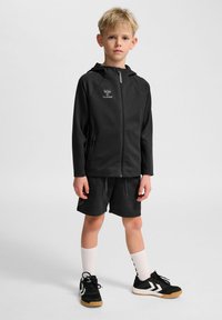Svart sportjacka med huva, svarta shorts, vita knästrumpor och svarta sneakers med vita detaljer. Innehåller dragkedja och logotypdetalj.