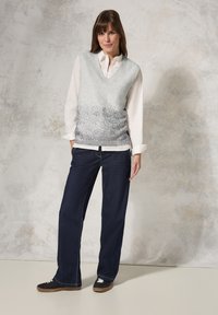 Suéter sin mangas con escote en V y un degradado de color plata a gris, combinado con una camisa blanca de manga larga y pantalones de mezclilla oscuros. Zapatillas negras.