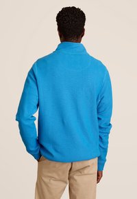 Blauwe gebreide trui met een omgevouwen kraag en lange mouwen, met een zachte textuur. Gedragen over beige broeken, gezien van de achterkant.