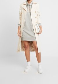Trench beige con bottoni dorati sopra un vestito felpa grigio. Sneakers bianche e una piccola borsa bianca completano l'outfit. Texture liscia.