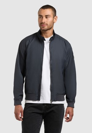 Man wearing a dark navy zip-up jacket over a white t-shirt and dark jeans, standing against a gray background, looking left.