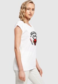 Camiseta de algodón blanca con un gráfico en rojo y negro del "Hellfire Club" que presenta un diseño de diablillo y espadas cruzadas. Mangas cortas, corte relajado.