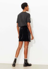 Chemise noire à manches courtes, associée à une mini-jupe plissée en velours noir. Le mannequin porte des bottines Chelsea noires avec des chaussettes grises.