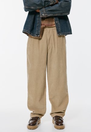 Beige Corduroy-Hosen mit weitem Bein, strukturiertem Material und elastischem Bund, kombiniert mit dunkelbraunen Schnürschuhen. Darüber eine Jeansjacke.