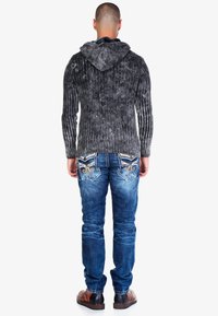 Graue gerippte Kapuzenjacke aus strukturiertem Stoff, getragen mit blauen verwaschenen Jeans mit dekorativen Gesäßtaschen und braunen Schuhen.