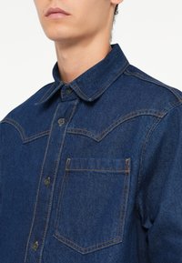 Camicia di jeans di colore blu scuro con colletto classico, chiusura frontale con bottoni e una singola tasca sul petto. Presenta cuciture a contrasto e una finitura testurizzata.