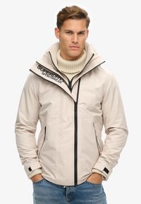 Beige jakke med høj krave, lynlåslukning foran og sorte detaljer. Har logo på skulderen og teksturerede manchetter. Bæres over en cremeløs sweater.