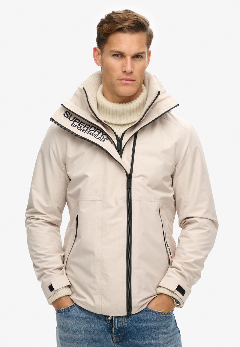 Beige jakke med høj krave, lynlåslukning foran og sorte detaljer. Har logo på skulderen og teksturerede manchetter. Bæres over en cremeløs sweater.