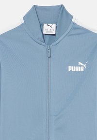 Veste zippée bleu clair avec un col côtelé, présentant des accents latéraux blancs et un logo Puma blanc sur la poitrine gauche. Texture de tissu lisse.